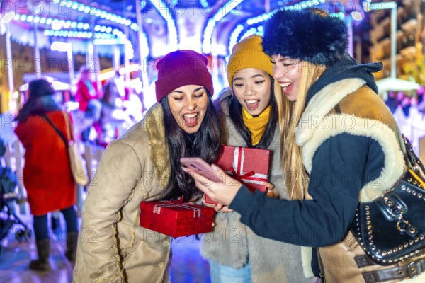 Three cheerful young women holding christmas gifts and using a smartphone in a christmas market with illuminated carousel in the background