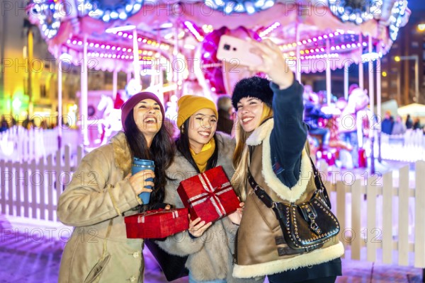 Three cheerful young women taking a selfie at a christmas market, holding gifts and warm drinks, with a carousel illuminated in the background