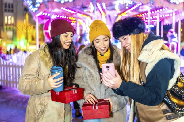 Three happy women using a smartphone and holding christmas gifts at night in a christmas market with a carousel in the background