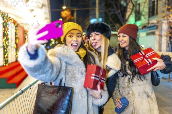 Three happy female friends taking a selfie while holding christmas gifts, enjoying a night out at a decorated christmas market