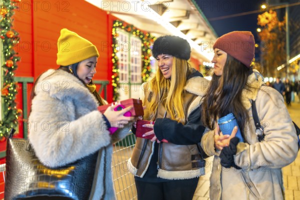 Three cheerful young women exchanging christmas presents at a decorated outdoor night market, enjoying the festive atmosphere