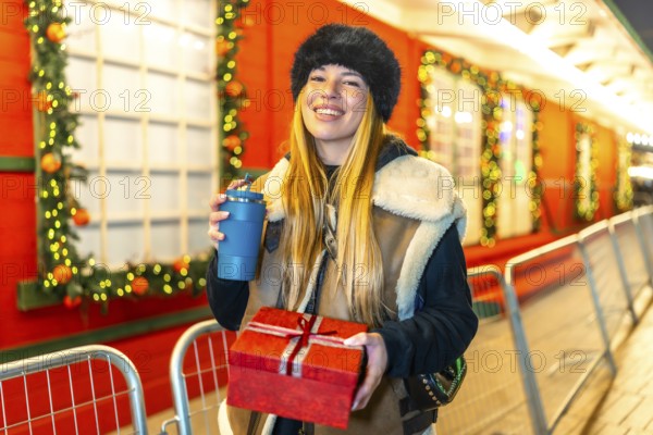 Young woman enjoying christmas spirit at a decorated outdoor market, holding a christmas present and a reusable cup