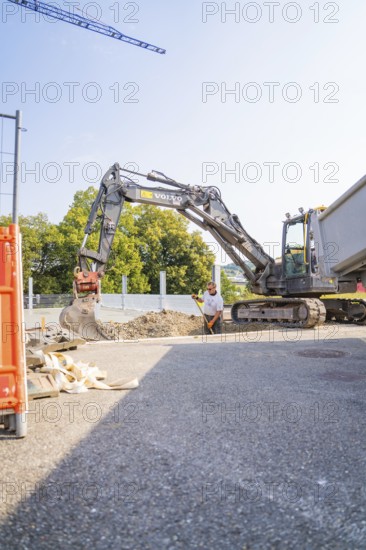 A large excavator and a construction worker on a building site with gravel and a lorry. Blue sky and trees in the background, car park construction, Calw, Germany
