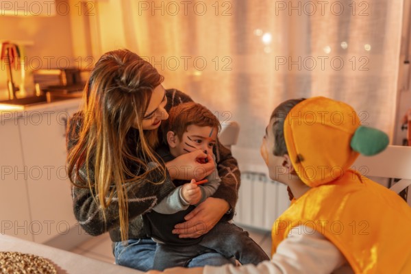 Mom applying halloween makeup on her son's face while his brother, dressed in a pumpkin costume, watches with amusement in a brightly lit kitchen