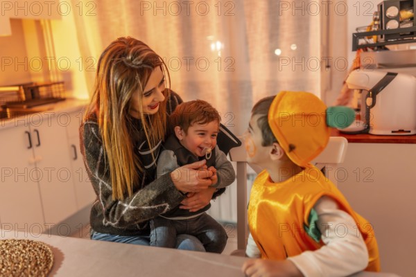 Mother holding baby while older brother wearing pumpkin costume interacting during halloween celebration at home