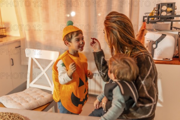 Mother applying halloween makeup to her son wearing a pumpkin costume while holding her baby in the kitchen