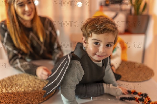 Smiling toddler wearing a bat costume crawling on a table while getting ready for a halloween party at home with his mother
