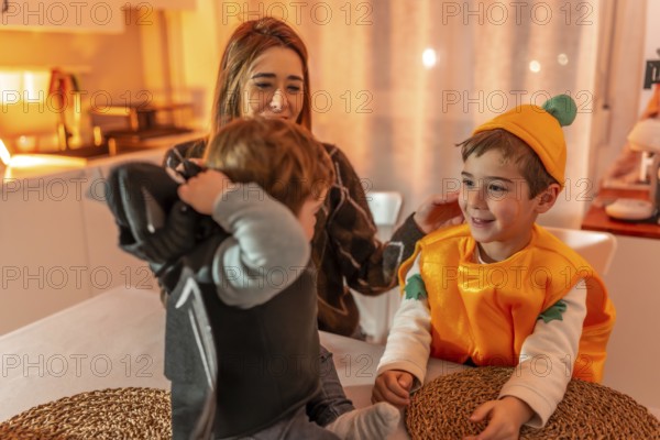 Happy family preparing costumes for halloween party at home, mother helping children wearing bat and pumpkin disguise