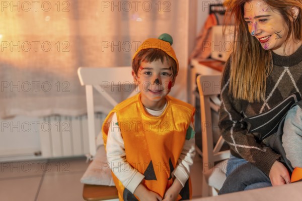 Happy child wearing pumpkin costume smiling and looking away while sitting next to his mother during halloween party at home