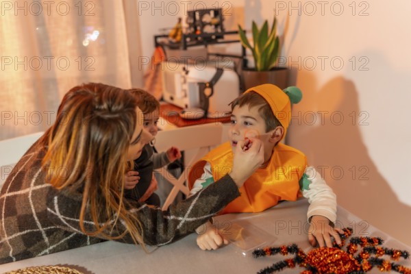 Mother putting halloween makeup on her son, who is wearing a pumpkin costume, while holding her baby in her arms, at home