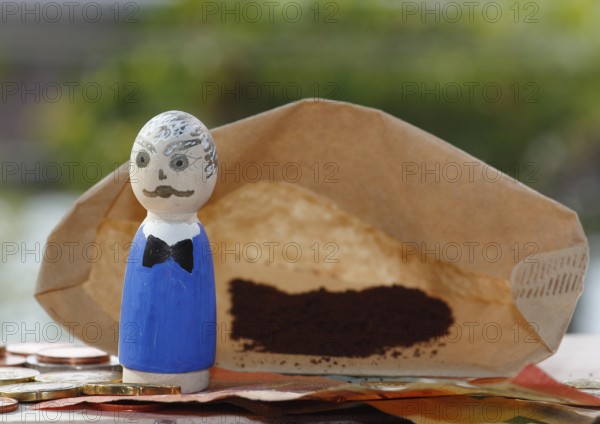 Male wooden cone figure with coins and banknote in front of filter bag with coffee North Rhine-Westphalia, Germany