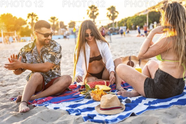 Three friends relaxing on a beach towel, sharing fresh fruit like watermelon and pineapple while enjoying a stunning sunset together