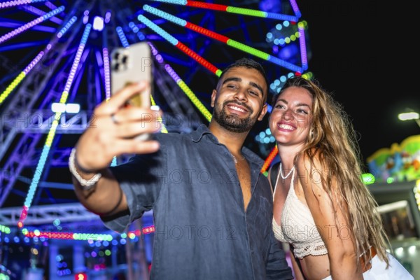Young couple smiling and taking a selfie with smartphone in front of a colorful ferris wheel at an amusement park at night