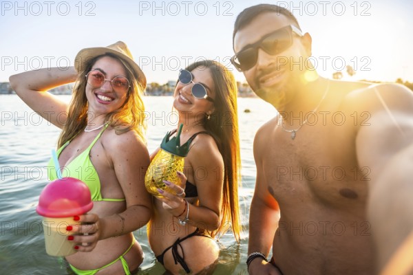 Three cheerful friends are taking a selfie in the sea, holding refreshing cocktails, enjoying a beautiful sunset during their summer vacation