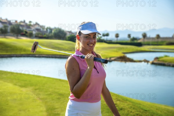 Female golfer smiling and holding her putter on shoulders on a sunny day on the golf course next to a water hazard