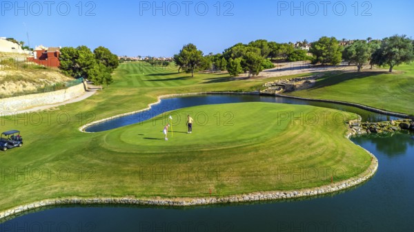 Two golfers are putting on a green surrounded by water, with lush fairways and trees in the background on a sunny day