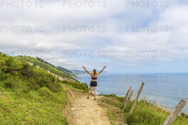 Woman hiker celebrating scenic coastal view with raised arms at sakoneta beach flysch cliffs in zumaia, gipuzkoa, basque country, spain