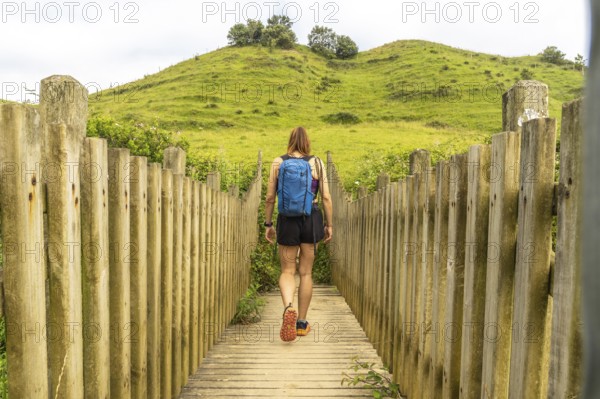 Female hiker with backpack walking on wooden footbridge leading towards a green hill in zumaia, basque country, spain
