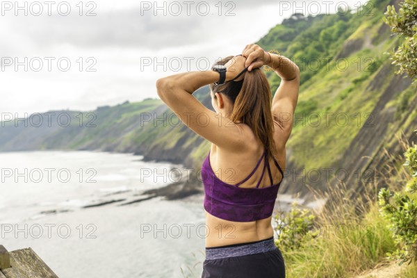 Rear view of a sportswoman tying her ponytail while enjoying the view of the sea and the flysch cliffs at sakoneta beach in zumaia, basque country, spain
