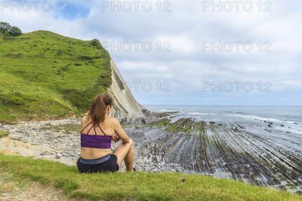 Woman sitting on green grass overlooking the impressive flysch cliffs and coastline of sakoneta beach in zumaia, gipuzkoa, basque country