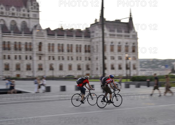 Two tourists riding bicycles in front of the hungarian parliament building in budapest, hungary