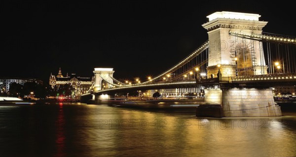 Chain bridge reflecting on danube river with gresham palace in background at night, budapest, hungary