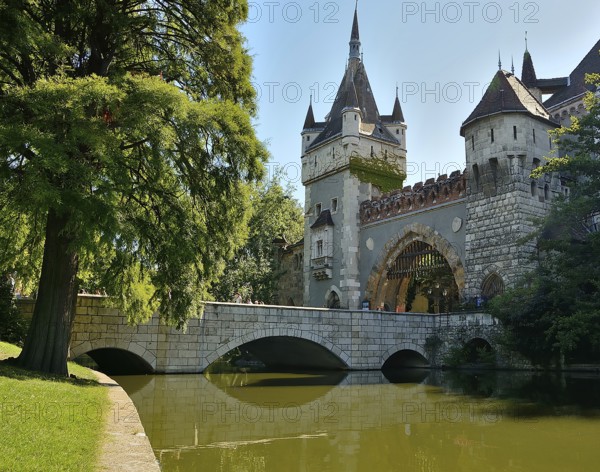 Tourists walking over stone bridge leading to vajdahunyad castle reflecting on lake in budapest, hungary