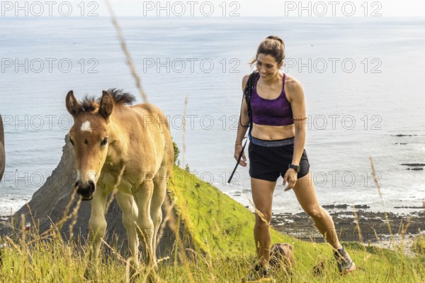 Female hiker with her dog encountering wild horses while walking on the cliffs of sakoneta beach, next to the flysch of zumaia, basque country, spain