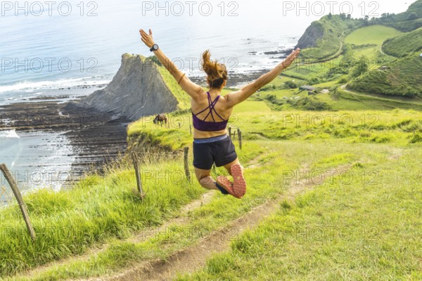 Sportswoman celebrates achieving a fitness goal by leaping in the air with arms outstretched, enjoying the breathtaking view of sakoneta beach and the flysch cliffs in zumaia, basque country