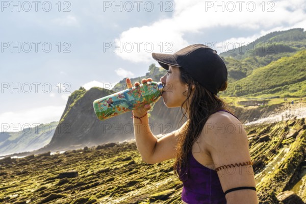Female athlete hydrating after exercising on sakoneta beach, enjoying breathtaking views of zumaia flysch cliffs in basque country, spain