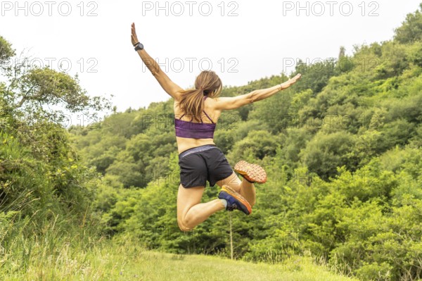 Celebrating athletic achievement, a woman jumps with outstretched arms amidst the verdant hills near sakoneta beach and the flysch cliffs of zumaia, gipuzkoa, in the basque country
