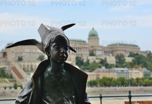 Bronze statue of a little princess wearing a bird shaped hat, overlooking the majestic buda castle on a sunny day in budapest, hungary