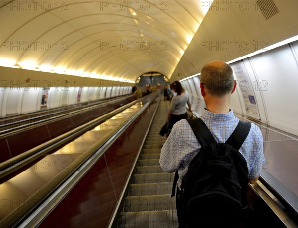 Tourist with a backpack ascending an escalator in a bustling subway station, surrounded by urban architecture and modern transportation