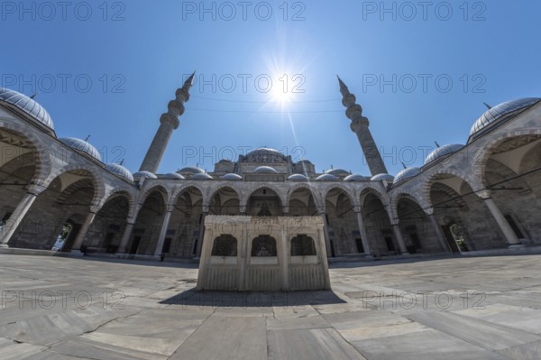 Wide angle view of the courtyard of suleymaniye mosque in istanbul, turkey, featuring intricate stonework, arches, domes, minarets, and the sun shining brightly overhead