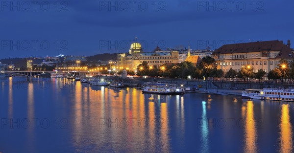 City lights reflecting in vltava river with illuminated buildings and boats moored along the embankment at dusk in prague