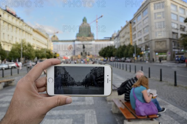 Hand holding smartphone taking photo of wenceslas square in prague, czech republic, with tourists sitting on bench in background