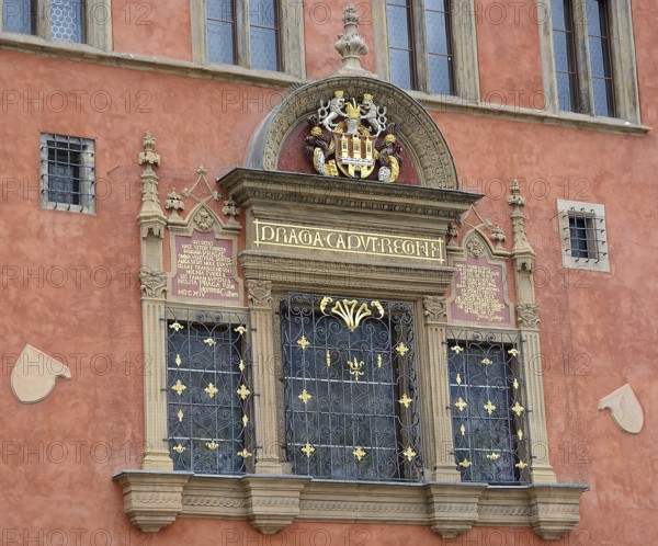 Architectural detail showcasing an ornate window with a coat of arms, latin inscription, and decorative ironwork on a vibrant, historic building in prague