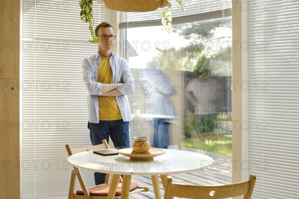 A middle-aged man with glasses stands with crossed arms beside a round table in a well-lit, modern living area. The natural light illuminates the space, creating a calm atmosphere