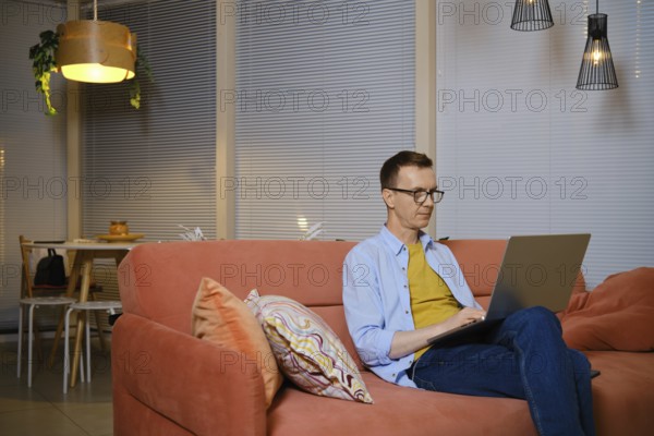 A man sits comfortably on a sofa and chats online on his laptop during evening hours. He communicates with enthusiasts from all over the world and shares a common hobby