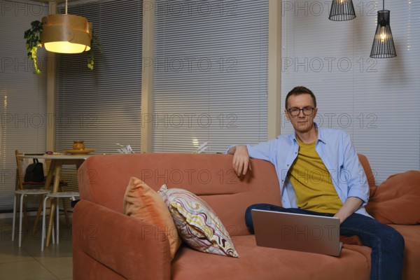 A man sits comfortably on a coral sofa, focused on his laptop in a cozy living room decorated with warm lighting. He is watching a new episode of the series online