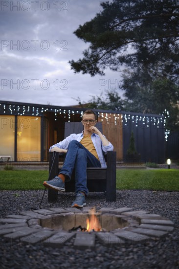 A person sits comfortably in a chair by a fire pit, enjoying a quiet evening outdoors. The setting features twinkling lights in the background, creating a cozy atmosphere in the backyard