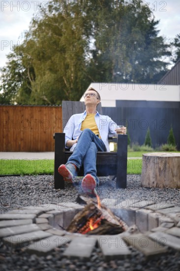 A middle-aged man enjoys a tranquil warm evening in his backyard, sitting in a wooden chair next to a fire pit. He sips a drink while gazing thoughtfully at the sky, surrounded by a peaceful landscape