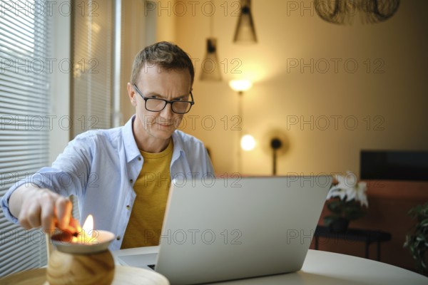 A middle-aged man lights small candle with lighter while working on his laptop at a round table in a warm, softly lit room in the evening
