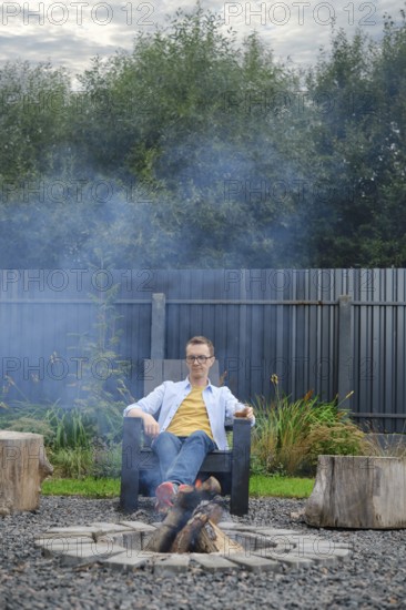 A middle-aged man sits comfortably in a chair next to a campfire surrounded by logs. Smoke rises gently in the cool air in the backyard. He looks thoughtfully at the flame and is lost in thought