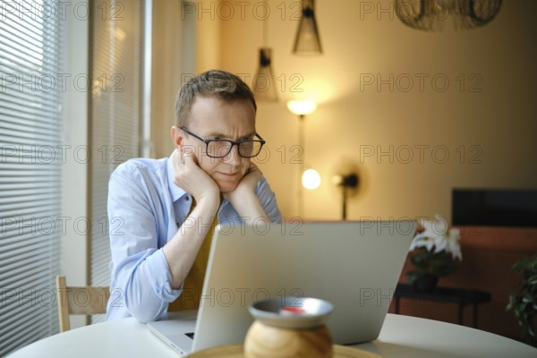A middle-aged man with glasses watching series on his laptop while seated at a round table in a stylish home. Natural light fills the space, creating a calming atmosphere for productivity