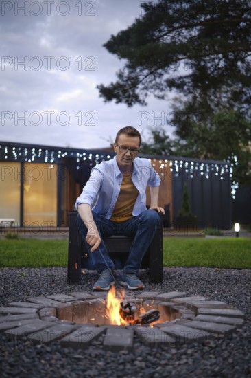 A man sits on a black chair, attentively tending to a fire pit surrounded by stones. The evening sky is grey, and twinkling lights illuminate a modern building in the background