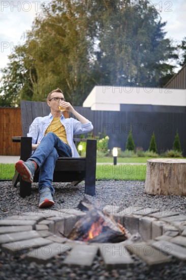 A middle-aged man sits in an outdoor chair, sipping a drink while enjoying the warmth of a fire pit. The serene backyard features lush greenery and a peaceful atmosphere, perfect for relaxation