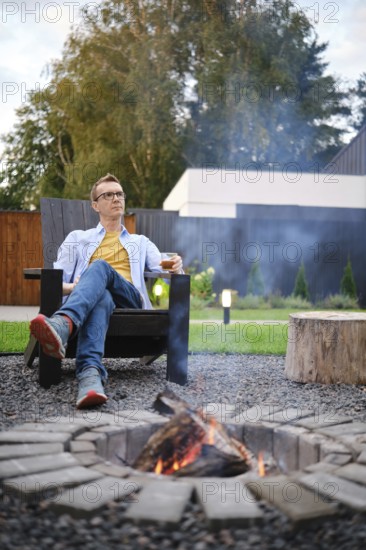 A retired man sits comfortably in a wooden chair by a crackling fire pit in his backyard. He enjoys a drink while surrounded by nature and greenery