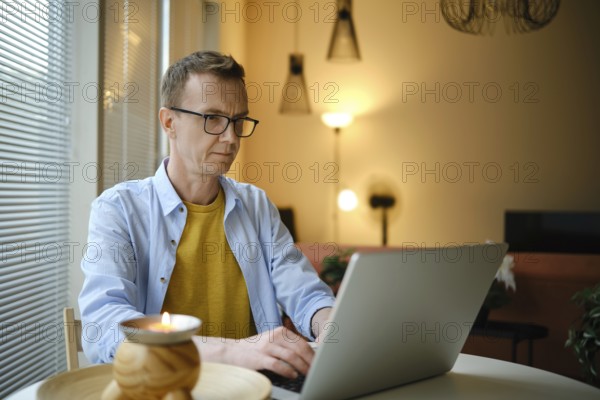 A middle-aged man sits at a table and typing letter on his laptop in a living room. Soft lighting and decor create a cozy atmosphere as he engages in his work