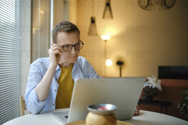 A focused man with glasses is working on his laptop with financial documents at home in the evening. He sits at a table surrounded by warm lighting and counting income and profit